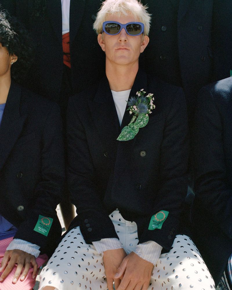 A group of fashionably dressed people, including a blonde adult male in blue sunglasses and a dark suit, pose outdoors under bright sunlight.