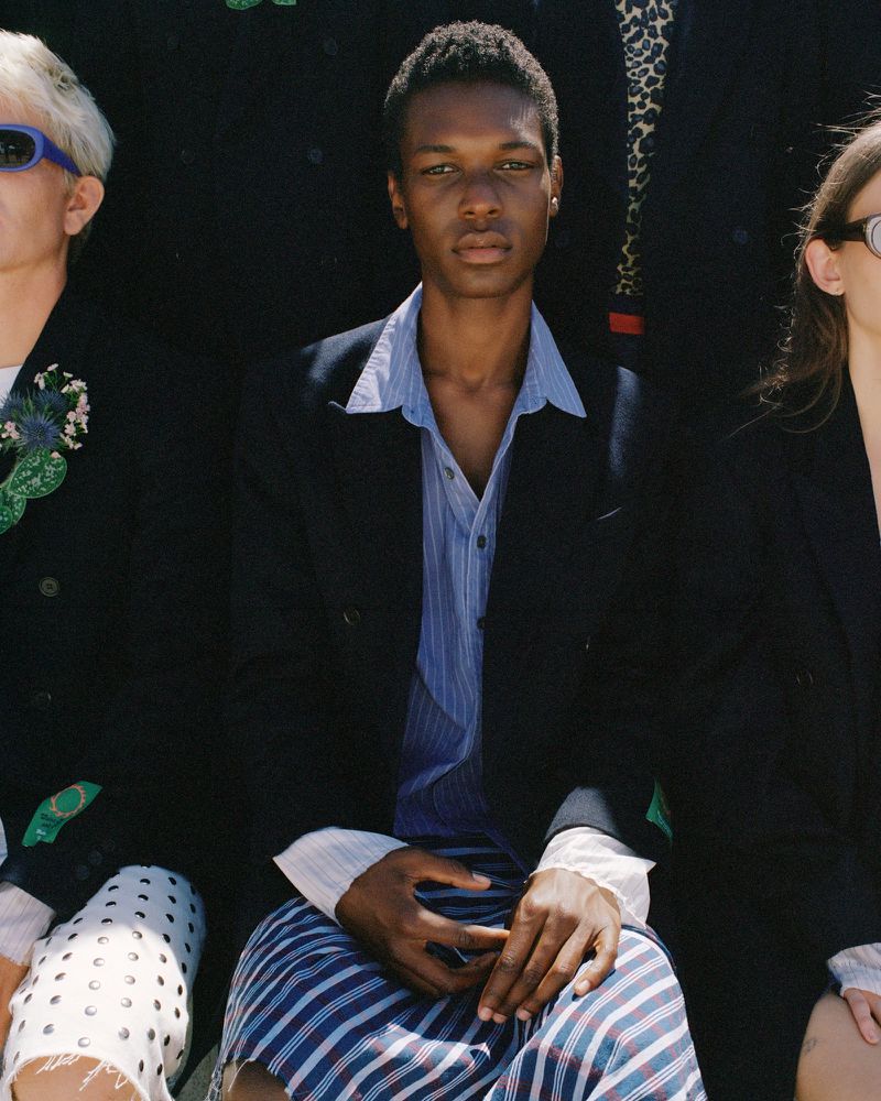 Editorial shot of a dark-skinned male model wearing a suit jacket and striped shorts, posed alongside other models in a dark studio.