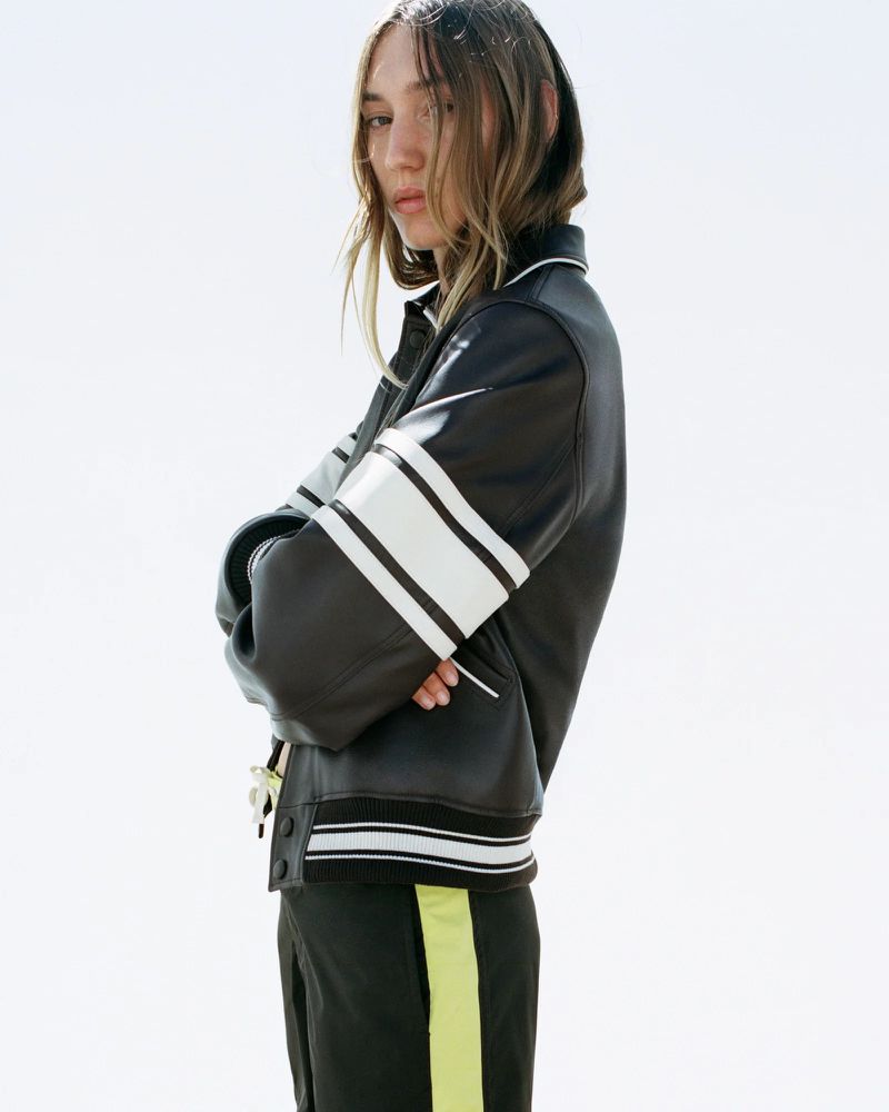 Young adult woman in a black jacket with white stripes stands against a bright sky, with her arms crossed in an outdoor setting.