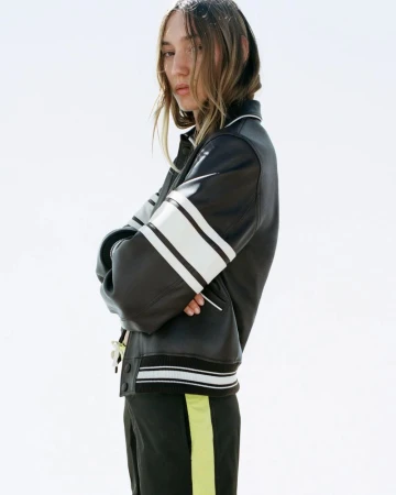 Young adult woman in a black jacket with white stripes stands against a bright sky, with her arms crossed in an outdoor setting.