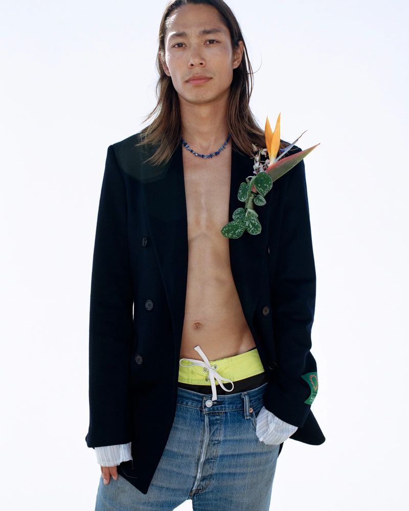 Young Asian man in a blazer, jeans and bare torso, standing in bright sunlight, accessorized with necklace and bird of paradise flower.