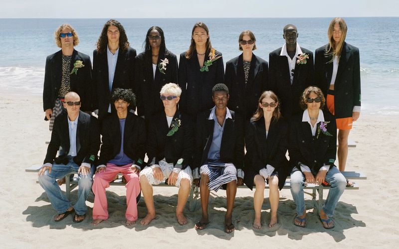 Group of people in blazers posing on a beach, with the ocean and sky in the background, under bright sunlight, fashion-forward style.