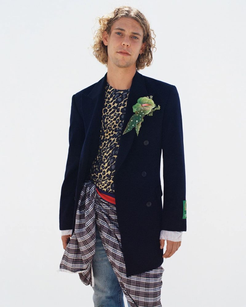 Man with blond hair poses in a navy blazer over a patterned t-shirt and jeans, wearing a decorative green floral pin.