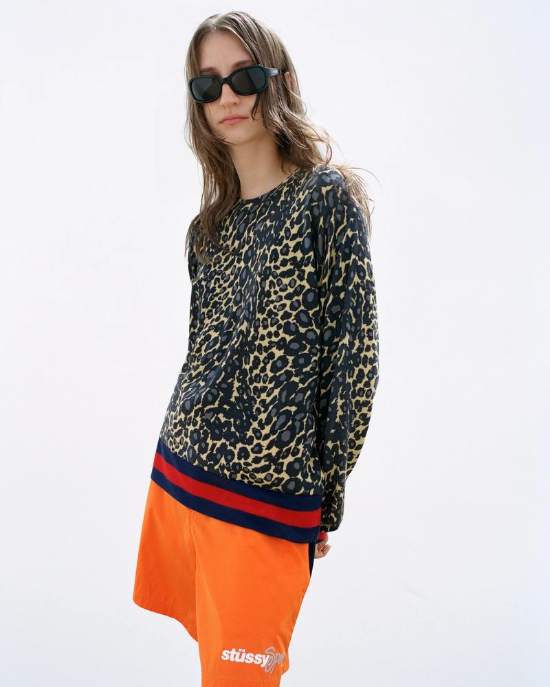 Woman in leopard print sweater and orange shorts poses in a bright white studio wearing dark sunglasses in a medium full shot.