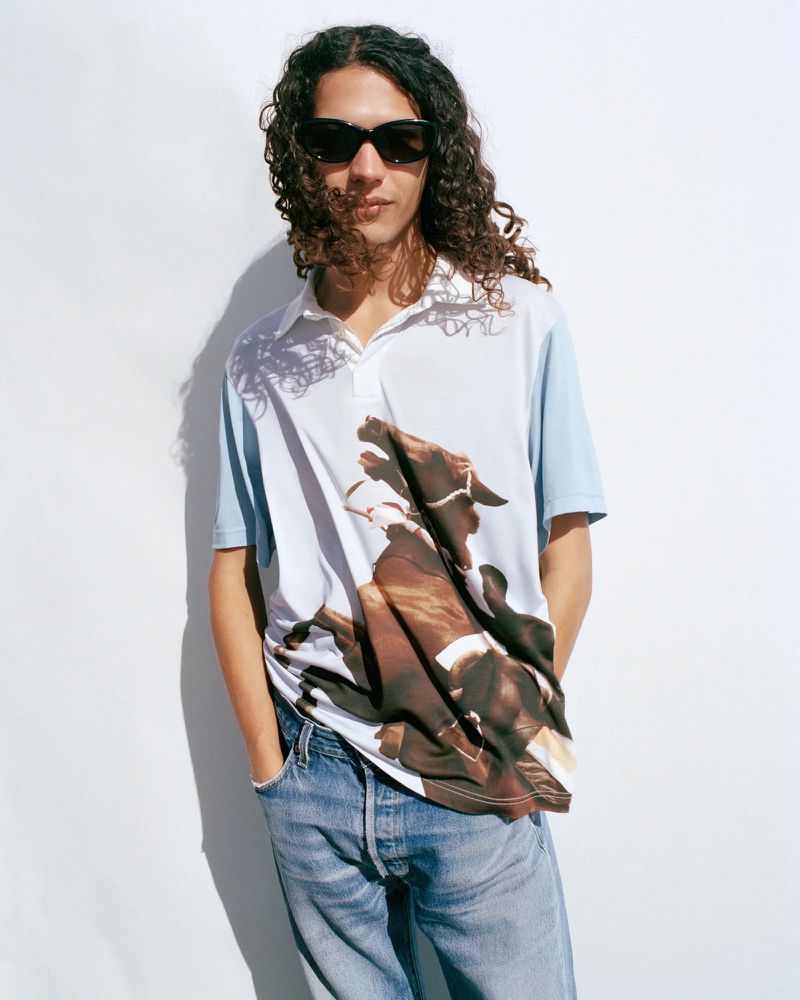 Young adult male with curly hair wears a polo shirt with a horse graphic, light blue jeans, and sunglasses against a white wall.