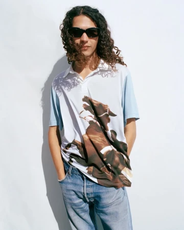 Young adult male with curly hair wears a polo shirt with a horse graphic, light blue jeans, and sunglasses against a white wall.