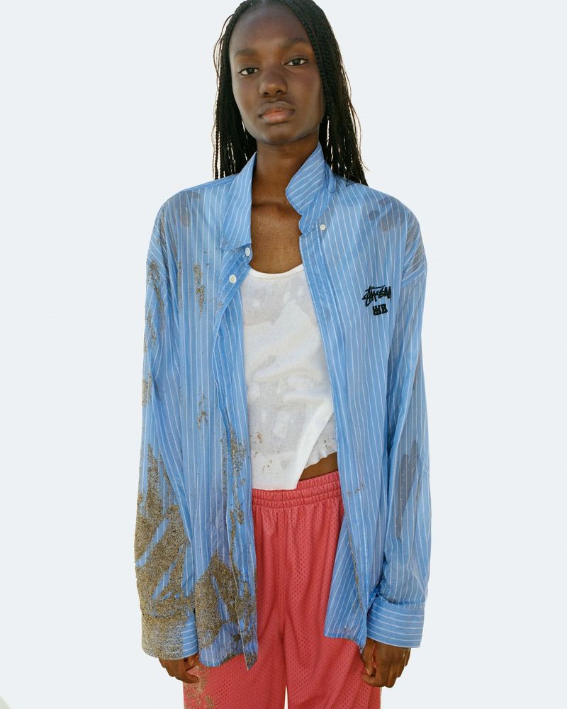 A young Black female model poses in an open blue striped shirt over a white tank top and pink shorts against a light backdrop.