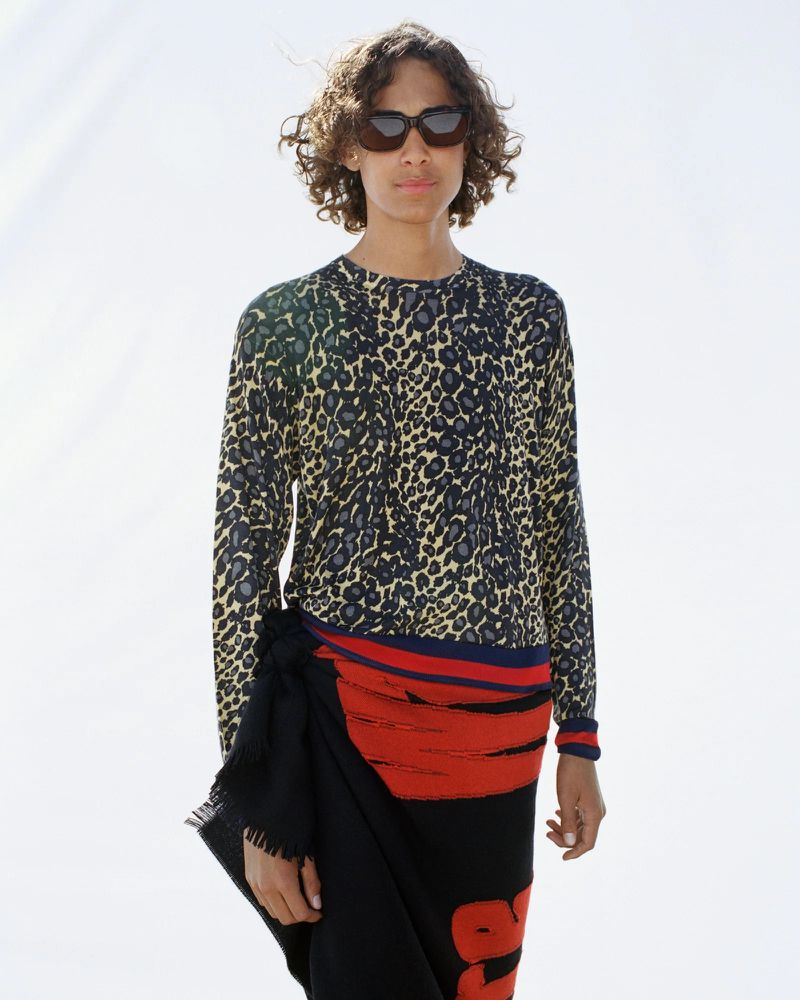 Fashion shot of a young adult with curly brown hair wearing sunglasses, a patterned long-sleeve shirt, and a black skirt against a bright background.