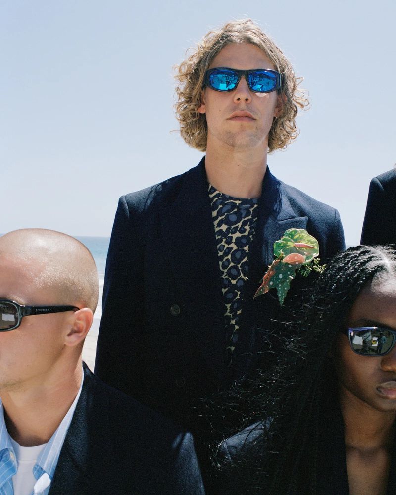 Group of three people in formal attire with sunglasses stand on a sunny beach with a clear sky, stylish and poised.