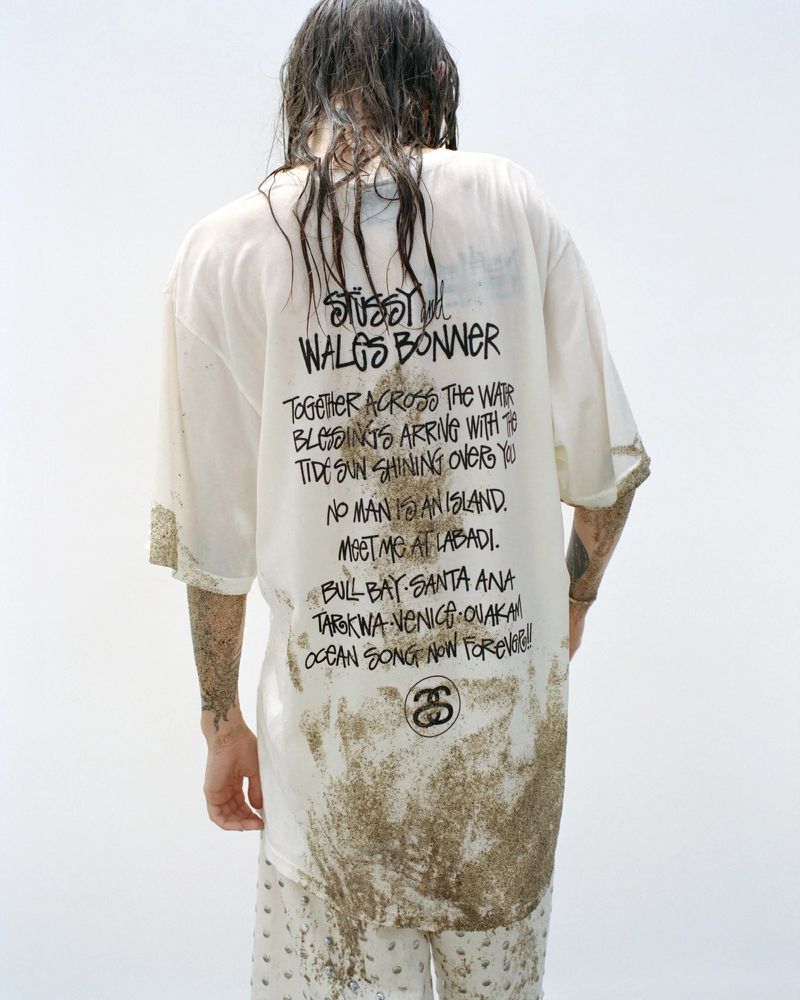 A person with wet hair wears a Stüssy Wales Bonner graphic tee and patterned pants against a bright background in a medium shot.