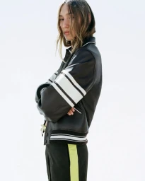 Ava Hawk McDean poses in profile wearing a black leather jacket with white sleeve stripes against a white studio backdrop.