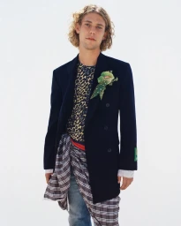 Hank Korsan in a navy double-breasted coat, leopard print top, and plaid waist wrap against a white studio background.