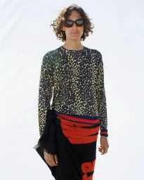 Model in dark sunglasses and a leopard print long-sleeve shirt with a black and red patterned wrap against a white studio backdrop.