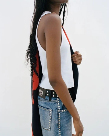 Side profile of a model wearing a white tank top and studded denim jeans with a logo belt against a plain white studio background.