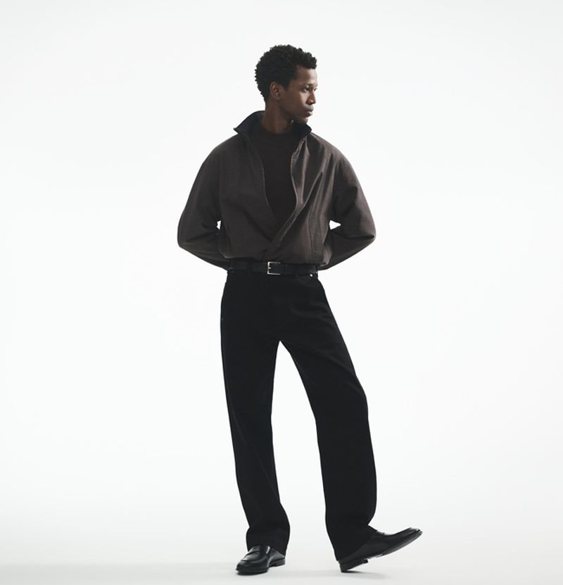A solitary Black male model poses in a minimalist studio setting, wearing a casual brown shirt, black pants, and dress shoes in a full shot.