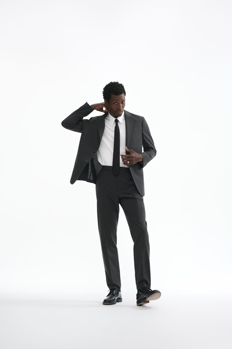 Calvin Royal III poses in a dark tailored suit, white dress shirt, and black necktie against a plain white studio background.