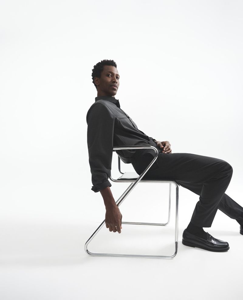 Black man poses seated on a metal chair in a photo studio wearing a black shirt, black pants, and black loafers against a white backdrop.