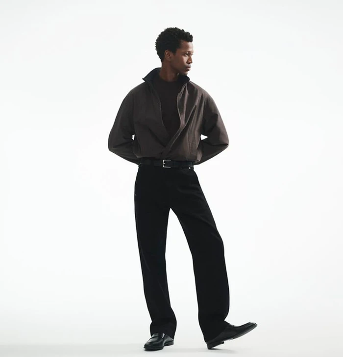 Calvin Royal III standing in a brown zipped jacket, black trousers, and black dress shoes against a white studio background.