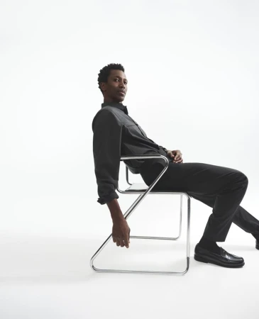 Calvin Royal III seated on a chrome cantilever chair wearing a dark grey shirt and black trousers against a white background.