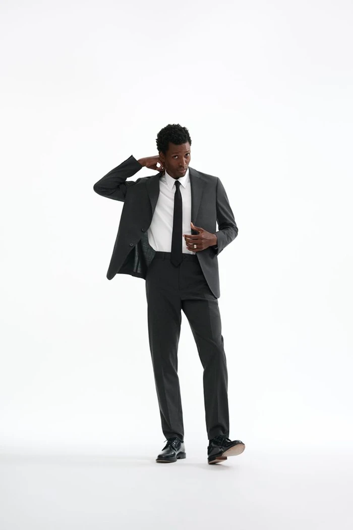 Calvin Royal III poses in a dark tailored suit, white dress shirt, and black necktie against a plain white studio background.
