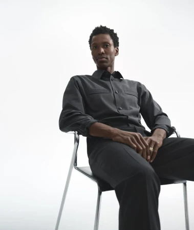 Calvin Royal III seated on a metal-framed chair wearing a dark grey button-up shirt and trousers against a white seamless background.