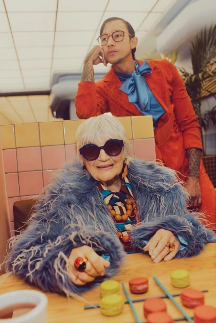 Elderly woman in a blue fur coat playing tic-tac-toe with macarons, while a young man in an orange suit looks on in an indoor setting.