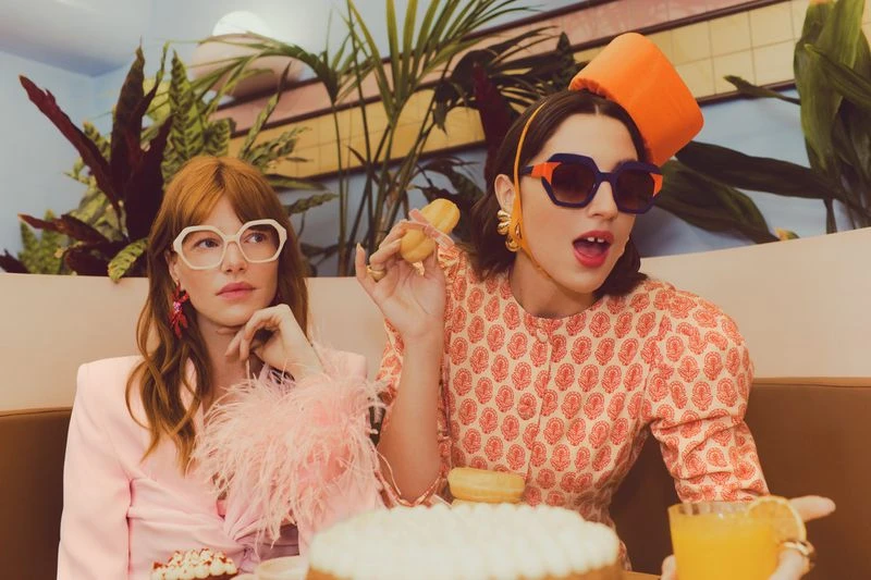 Two women in retro fashion eat pastries and drink juice indoors, featuring patterned dresses, sunglasses, hats, and decorative plants.