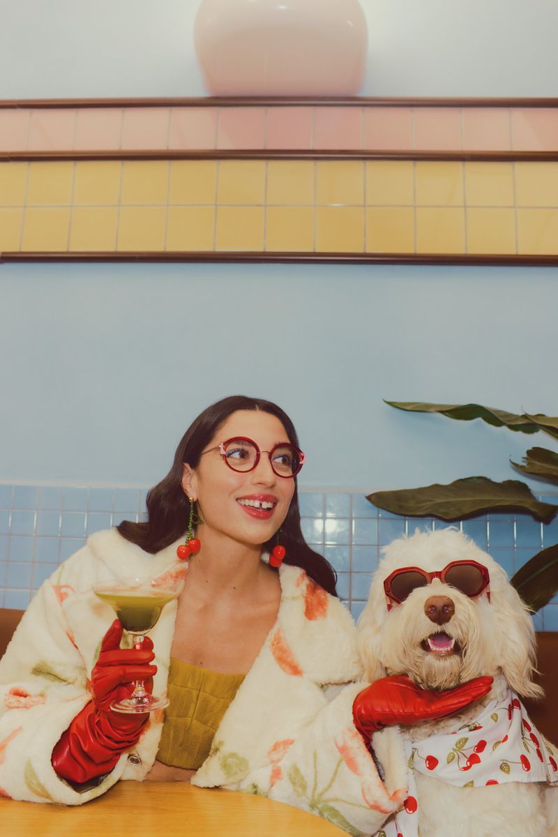 A woman in a floral robe and glasses poses with a dog wearing sunglasses and a cherry-print shirt while seated at a table indoors.