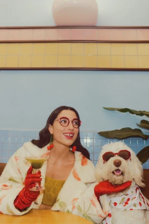 Woman in red eyeglasses and a plush coat holding a cocktail beside a white dog wearing sunglasses and a cherry bandana.