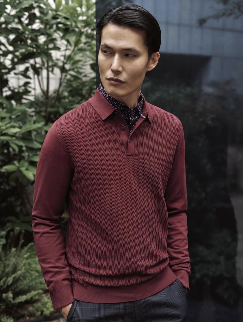 An Asian adult male in a maroon long-sleeve polo shirt stands against a background of green foliage, looking off to the side.