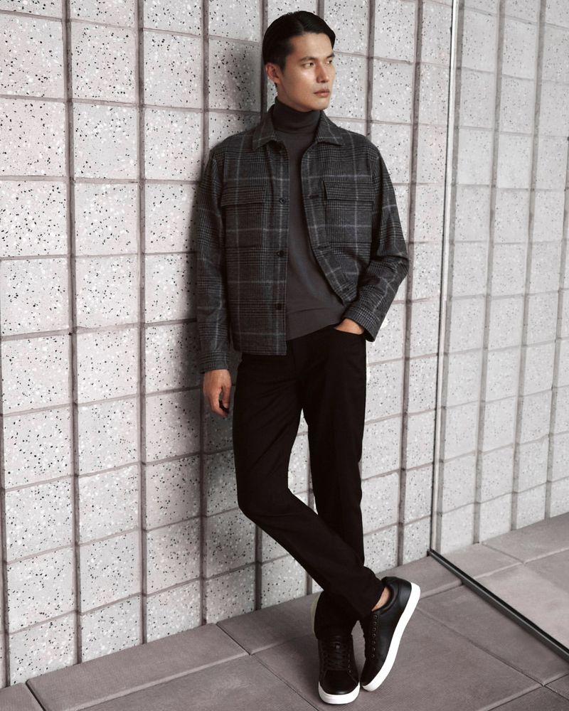 Stylish adult male wearing a grey checked overshirt, black turtleneck, slim black jeans, and sneakers leaning against a tiled wall.