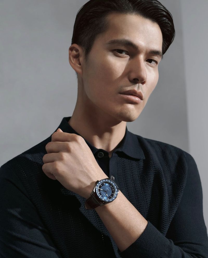 Close up of a handsome man wearing a navy sweater and a blue wristwatch with a brown leather band, set against a neutral grey background.