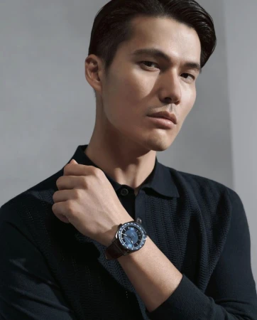 Close up of a handsome man wearing a navy sweater and a blue wristwatch with a brown leather band, set against a neutral grey background.