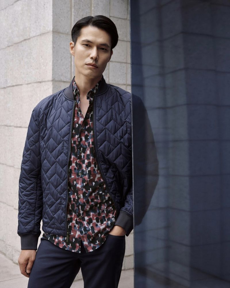 Portrait of a stylish Asian man in a quilted bomber jacket and patterned shirt, standing against a textured wall with a reflective blue panel.