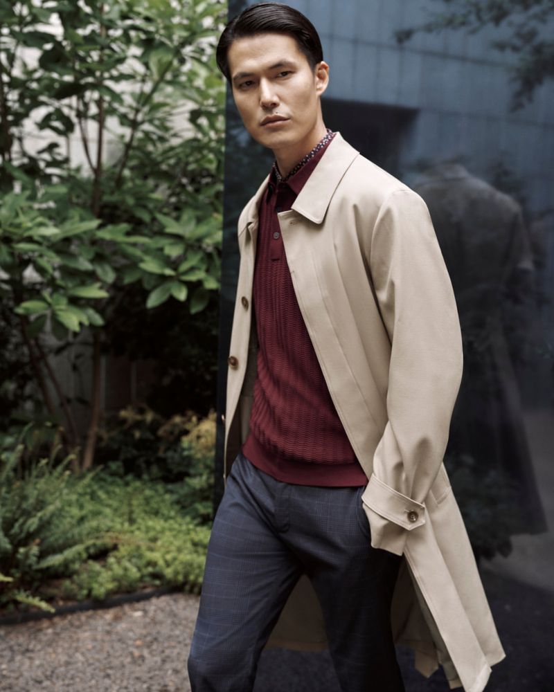 Portrait of an adult male posing outdoors in a beige overcoat, a dark-red patterned shirt, and dark pants against a leafy background.