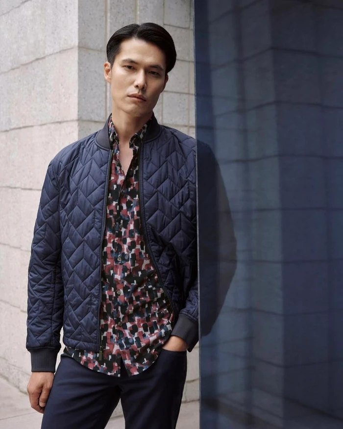 Model Dae Na wearing a quilted navy bomber jacket and patterned shirt against a stone exterior for Perry Ellis F/W 2025.