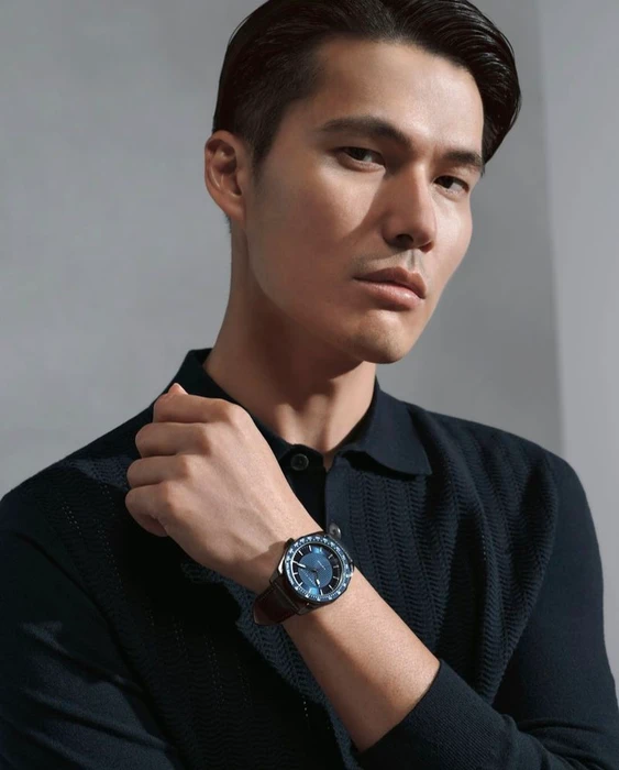 Model Dae Na wearing a navy textured polo and wristwatch for the Perry Ellis F/W 2025 campaign against a grey studio backdrop.