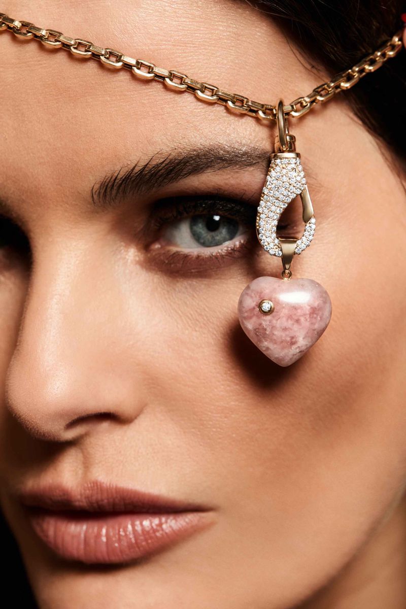 Close-up portrait of a woman with light skin wearing a gold chain headpiece with pink heart pendant, against a dark background.