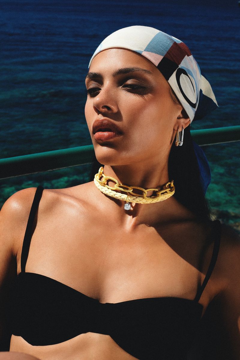 Woman wearing a headscarf and gold chain posing outdoors with a turquoise sea background, lit by natural sunlight. Beauty shot, fashion style.
