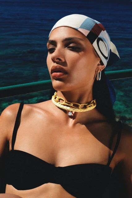 Woman wearing a headscarf and gold chain posing outdoors with a turquoise sea background, lit by natural sunlight. Beauty shot, fashion style.
