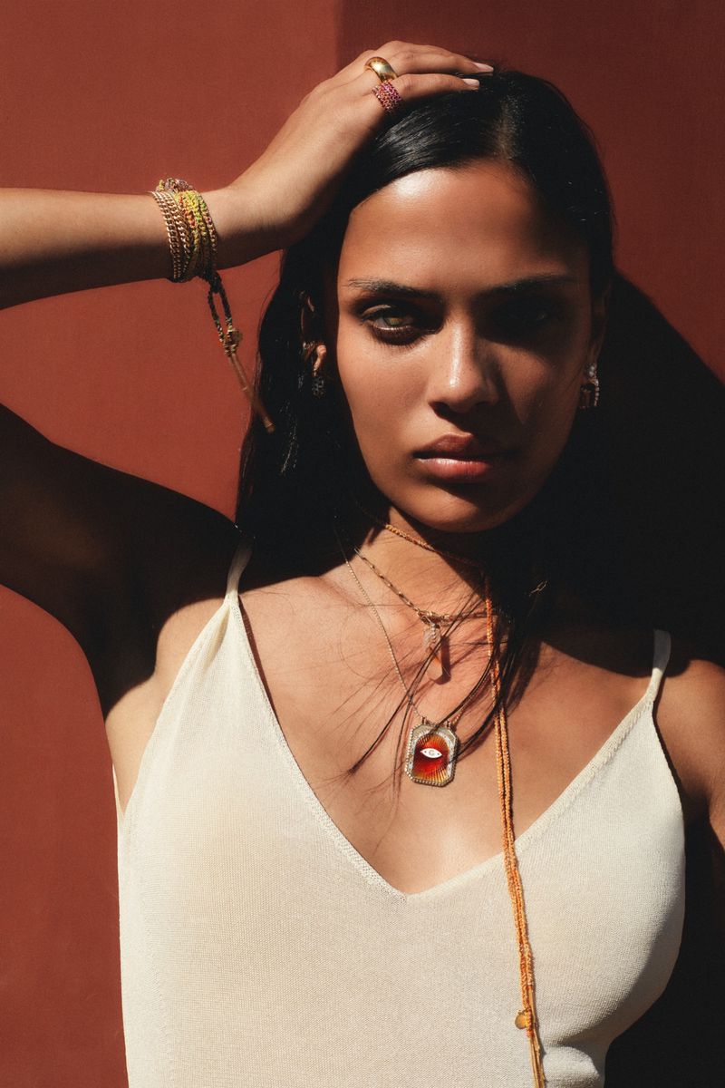 Close-up of a woman with dark hair wearing gold jewelry, posing in front of an orange background in soft, diffused lighting.