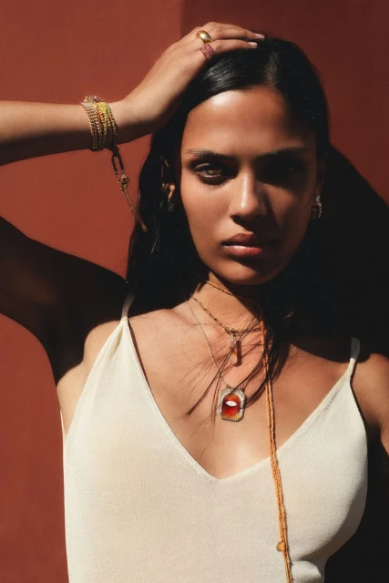 Close-up of a woman with dark hair wearing gold jewelry, posing in front of an orange background in soft, diffused lighting.