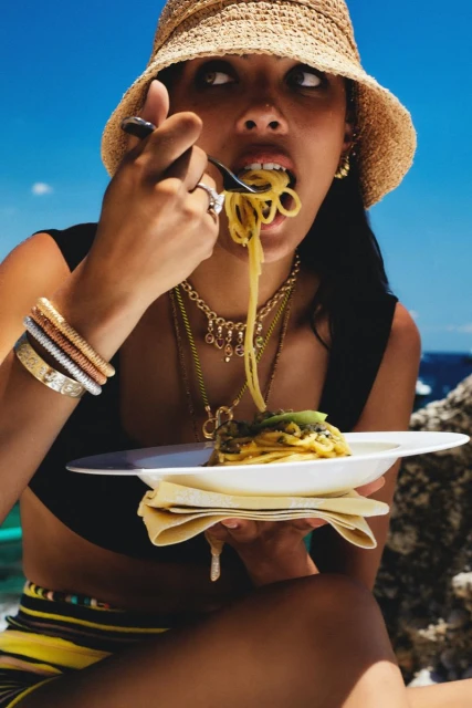 A young woman with dark hair and a straw hat eats spaghetti on a sunny day outdoors near a rocky coast with a turquoise sea.