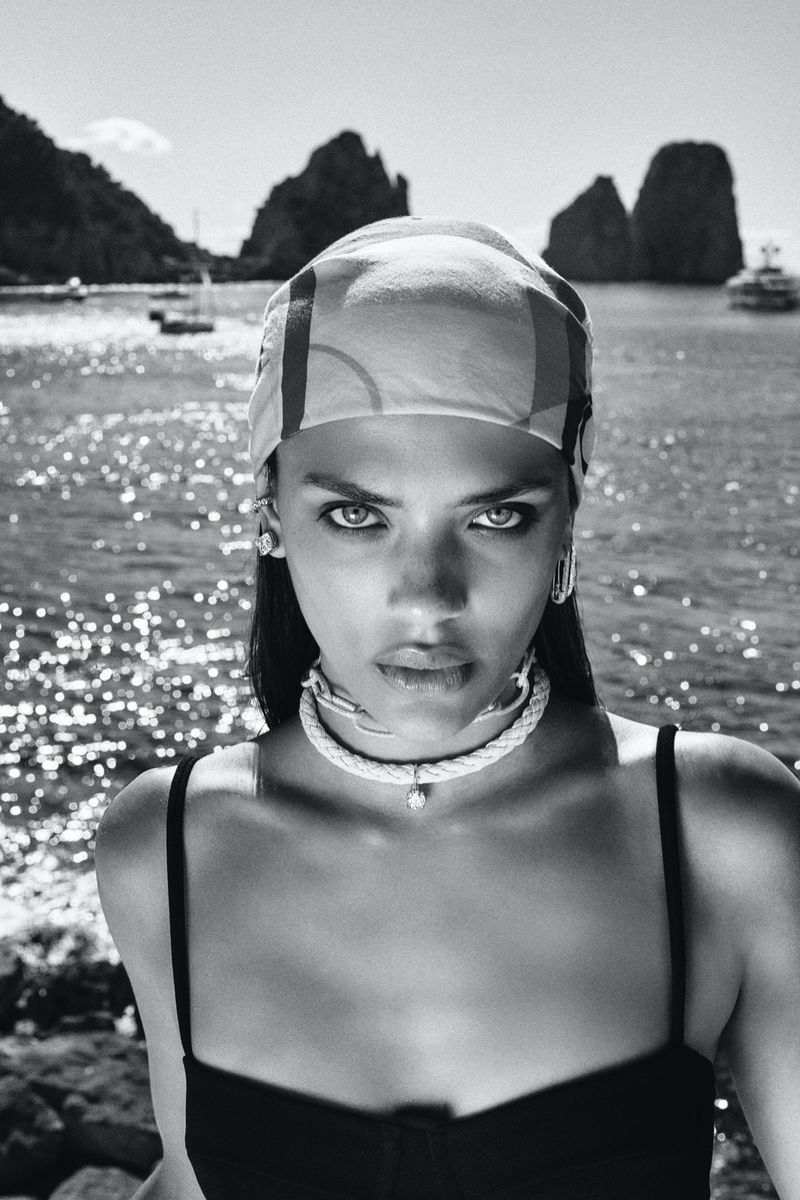 Black and white portrait of a woman wearing a headscarf, rope choker necklace, and black top against a sunlit coastal sea background.