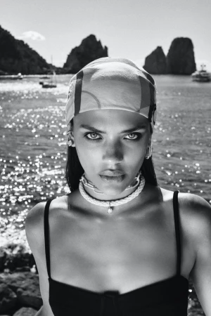 Black and white portrait of a woman wearing a headscarf, rope choker necklace, and black top against a sunlit coastal sea background.