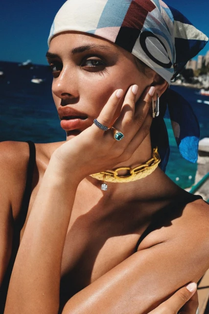Woman in headscarf and gold jewelry poses against an outdoor backdrop of the sea and sky on a sunny day in close-up.