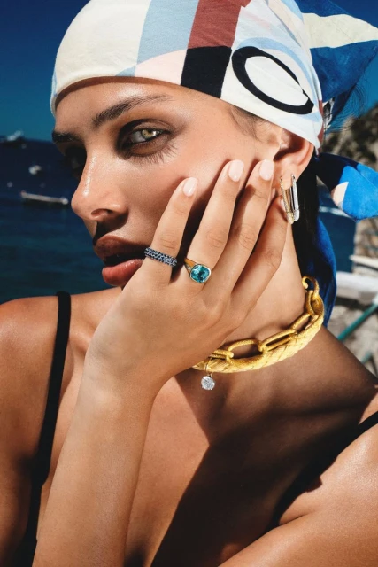 Close-up of a woman with a headscarf, jewelry, and tanned skin, posing outdoors with a blurry background of the sea and sky.