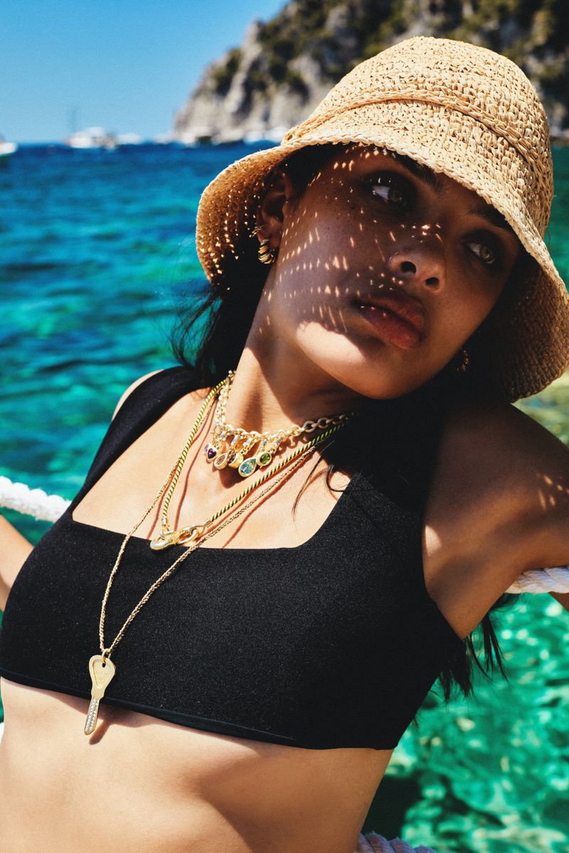 Brunette woman in black crop top and straw sun hat poses with necklaces on a boat with turquoise water and rocky cliffs behind.
