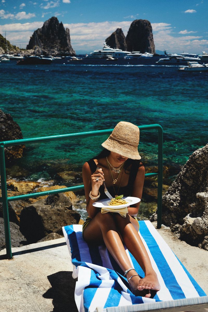 A tan-skinned adult female wearing a sunhat is relaxing on a beach chair while eating pasta with a scenic ocean view in the background.