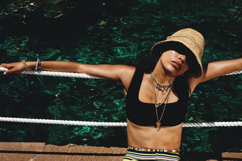 A woman in a straw hat poses near water, wearing a black top, striped skirt, and layered necklaces, leaning against a rope railing.